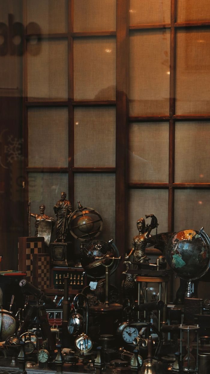A vintage shop window display featuring globes, clocks, and decorative statues with warm lighting.