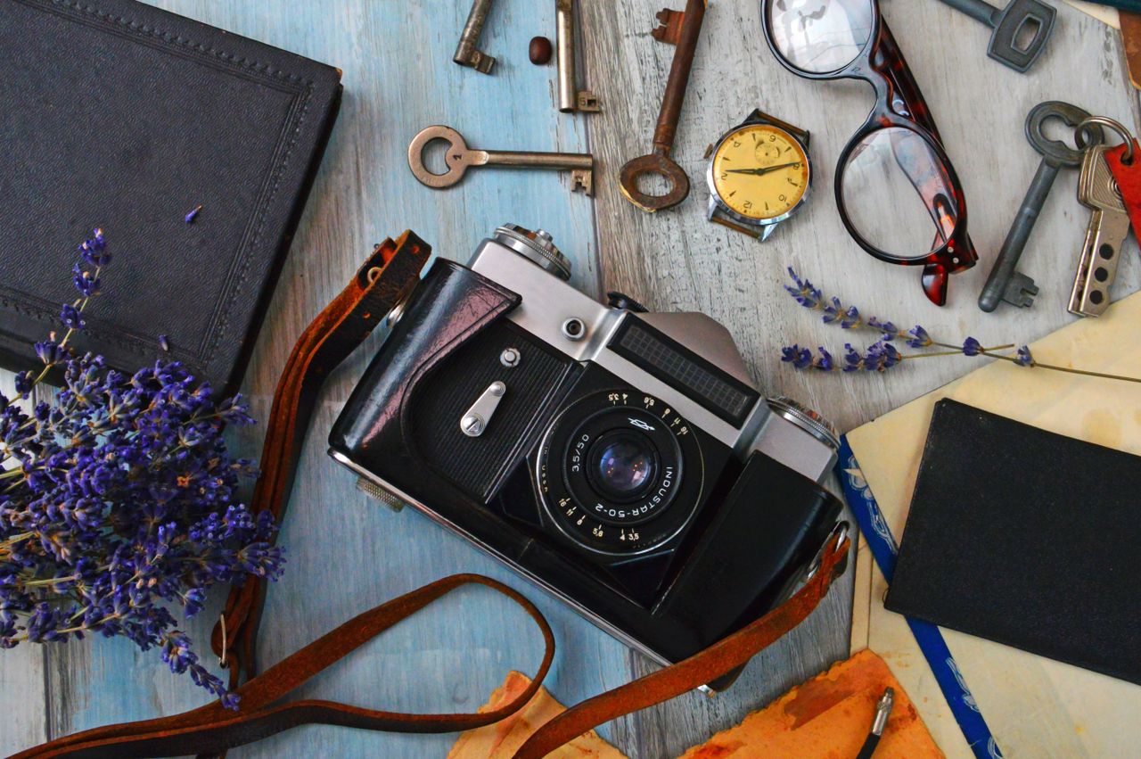 Flat lay of a vintage camera surrounded by keys, a watch, glasses, and lavender on a wooden table.